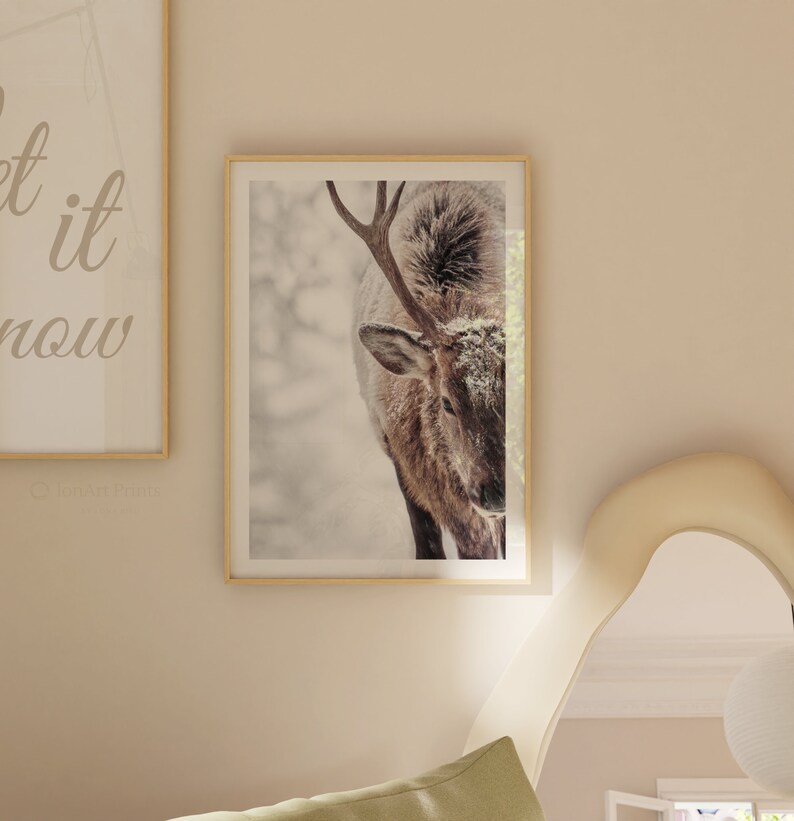 May include: A close-up of a brown and white deer with antlers standing in a snowy forest. The deer is looking down at the ground. The image is in a gold frame.