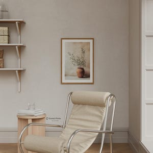 May include: A framed wall art print featuring a terracotta pot with green olive branches against a neutral background, displayed in a room with a beige armchair, a small wooden side table, and white wall-mounted shelves with books.