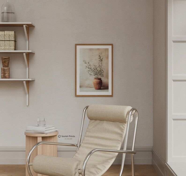May include: A framed wall art print featuring a terracotta pot with green olive branches against a neutral background, displayed in a room with a beige armchair, a small wooden side table, and white wall-mounted shelves with books.
