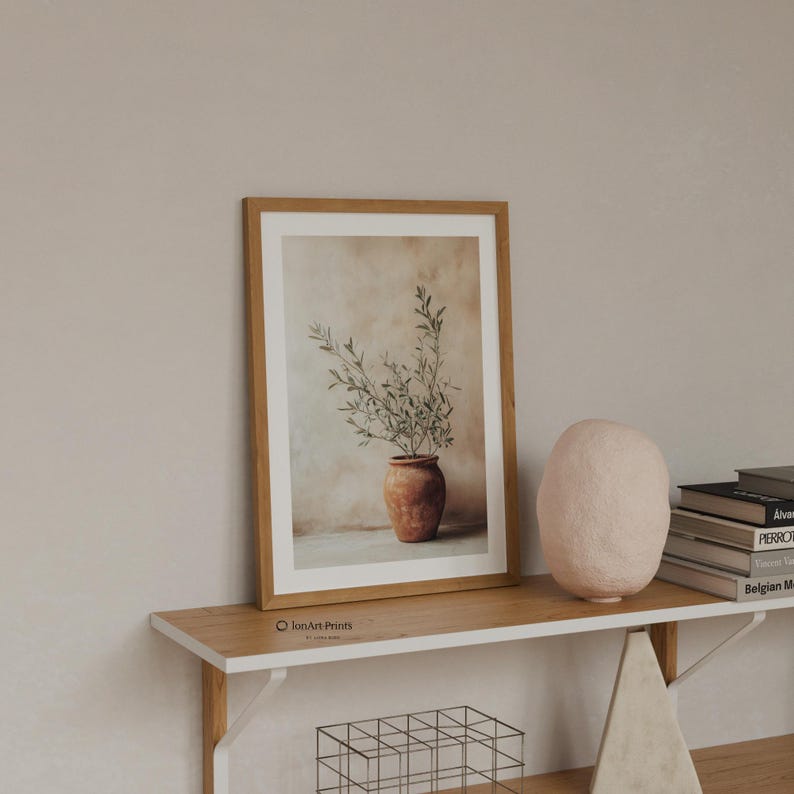 May include: A framed vertical wall art print featuring a terracotta pot containing an olive branch with green leaves against a beige background. The print is housed in a light wood frame and displays the text JonArtPrints in the bottom left corner.