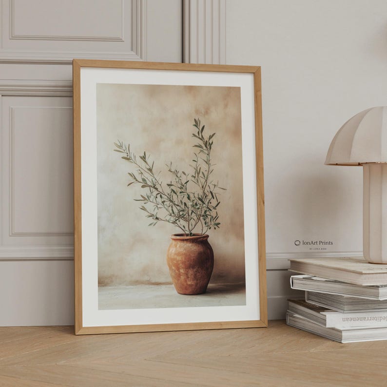 May include: A framed wall art print featuring a terracotta pot containing an olive branch with green leaves against a textured beige background. The print is housed in a light wood frame, with the text IonArt Prints visible on the lower right side.