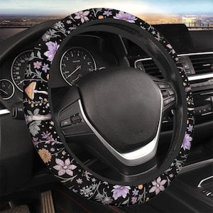 May include: Black steering wheel cover with a floral pattern featuring purple, pink, and yellow flowers, along with green leaves and mushrooms. The cover is set against a car's dashboard, with gauges and controls visible.