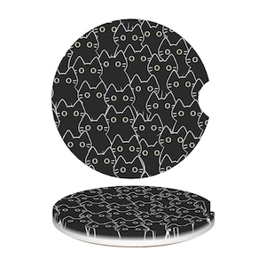May include: Two black car coasters with a repeating pattern of white-outlined cat faces with yellow eyes. One coaster has a notch for a car cup holder. The coasters are round and have a white border.