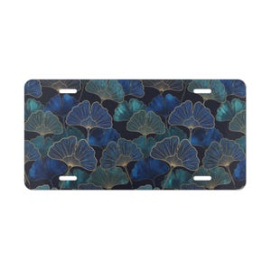 May include: License plate featuring a repeating pattern of stylized ginkgo leaves in shades of blue and teal with gold outlines. The background is a dark navy blue. The plate has four mounting holes.