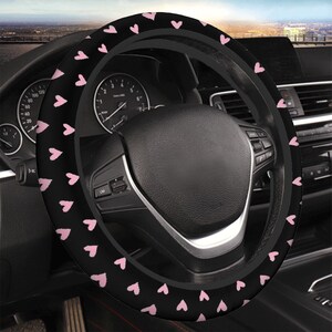 May include: Black steering wheel cover with a pattern of small pink hearts. The cover is shown installed on a car's steering wheel, with the dashboard and other interior details visible in the background.