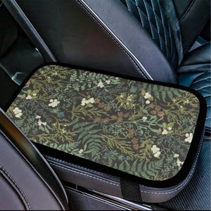 Puede incluir: Una funda rectangular para reposabrazos de coche con un fondo oscuro y un patrón botánico de helechos verdes, flores blancas y piñas marrones. Tiene un borde negro y una correa negra.