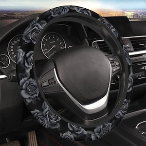 May include: Black steering wheel cover with a pattern of gray roses. The cover is installed on a car's steering wheel, with the dashboard and gauges visible in the background. The roses are detailed and create a striking contrast against the black.