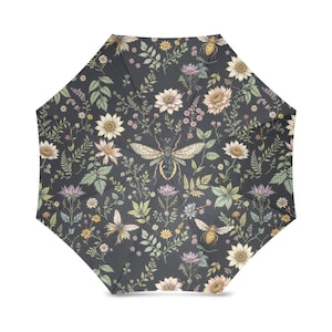 Puede incluir: Un paraguas abierto con un fondo gris oscuro, cubierto con un estampado floral con abejas y mariposas. El diseño incluye girasoles, margaritas y otras flores en tonos crema, amarillo y morado. El paraguas tiene forma octogonal.