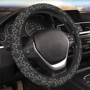 May include: Black steering wheel cover with a repeating pattern of white cat faces. The cover is fitted on a black steering wheel inside a car. The dashboard and other car components are visible in the background.