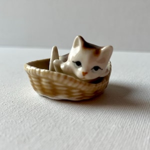 Może przedstawiać: Mała ceramiczna figurka kota szylkretowego spoczywającego w wiklinowym koszu. Kot ma białe, brązowe i płowe oznaczenia, czarne oczy i różowy nos. Kosz jest brązowy i ma plecioną fakturę.