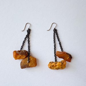 May include: Dangle earrings with rough amber stones in orange and brown hues. The amber pieces are suspended from dark metal chains and silver ear wires. A unique jewellery design with natural elements.