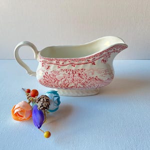 Op de afbeelding: Een witte keramische sausboot met een rood toile de Jouy-patroon. De boot heeft een handvat en een tuit. Decoratieve bloemen en een dennenappel staan op de voorgrond. Een klassiek stuk voor het serveren van sauzen.