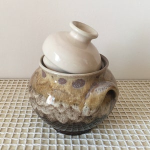 May include: A ceramic pot with a lid. The pot is a mix of brown, tan, and gray with a handle on the side. The lid is a light beige color and sits inside the pot. The pot is sitting on a white surface.
