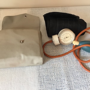 Può includere: Un misuratore di pressione sanguigna vintage con un quadrante bianco, un bracciale nero e una pera arancione e blu. Il misuratore è in una custodia di tela beige.