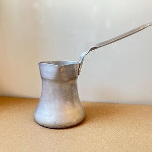 May include: A silver aluminum coffee pot with a long handle. The pot has a rounded base and a wide opening at the top.