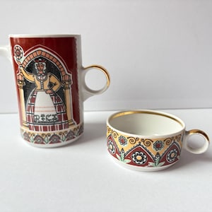 May include: A vintage ceramic creamer and cup set. The creamer is burgundy with a woman illustration and gold accents. The cup has a floral pattern with gold trim. Both pieces have a white interior and are on a white surface.