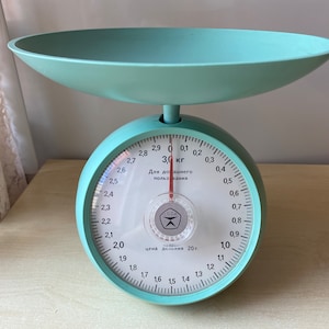 Op de afbeelding: Een vintage teal keukenweegschaal met een witte wijzerplaat en een rode naald. De wijzerplaat heeft markeringen voor kilogrammen en grammen. De weegschaal geeft "3,0 kg" en "Для домашнего пользования" (Voor huishoudelijk gebruik) aan.