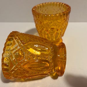 May include: Two amber-coloured, vintage-style glass cups. The cups have a detailed, textured pattern with diamond and oval shapes. One cup is upright, while the other is lying on its side, showcasing the intricate design.