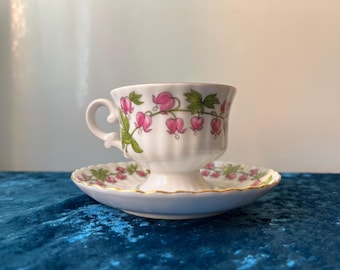 May include: A white teacup and saucer with a floral design. The cup and saucer have a gold trim and feature pink bleeding heart flowers and green leaves.