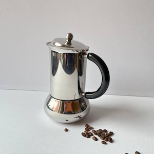 May include: A stainless steel stovetop espresso maker with a black handle and a small silver knob on the lid. The coffee maker is cylindrical with a rounded base. A small pile of coffee beans is next to the coffee maker.