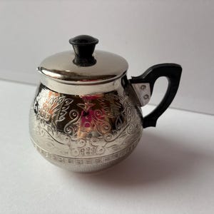 May include: A small, silver-colored teapot with a black handle and lid. The teapot has an ornate floral design etched into its surface. The handle and lid knob are black. The teapot is likely made of metal.