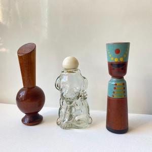 May include: Three vintage glass perfume bottles with different designs. The first bottle is a wooden stopper with a round base. The second bottle is shaped like a dog and has a white lid. The third bottle is a wooden figure painted with blue, red, and yellow details.