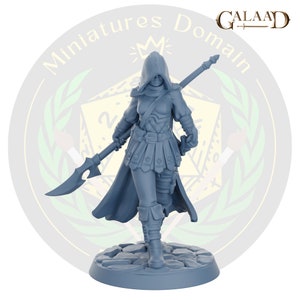 Op de afbeelding: Een grijs geverfd miniatuurbeeldje van een vrouwelijk personage dat een capuchonmantel draagt en een speer vasthoudt. Het personage staat op een ronde basis met een steenachtige textuur.