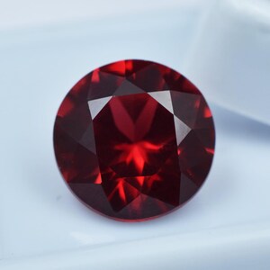 Può includere: Una gemma rotonda e sfaccettata di colore rosso intenso. La gemma presenta molte sfaccettature triangolari che riflettono la luce, creando una brillantezza. La pietra è posta su uno sfondo bianco, evidenziando il suo colore ricco e la sua chiarezza. Adatta per gioielli.