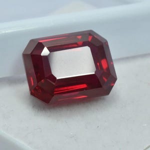 Peut inclure: Une pierre précieuse rouge foncé taillée en rectangle. La gemme présente une surface brillante et réfléchissante et un centre clair et ouvert. La taille comporte de multiples facettes qui captent et réfléchissent la lumière, améliorant l'éclat de la pierre. La pierre est posée sur un fond blanc.