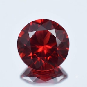 May include: A round, faceted gemstone with a deep, vibrant red color. The stone features a precise, symmetrical cut with multiple triangular and kite-shaped facets that create a brilliant, reflective surface and a clear, sharp geometric pattern.