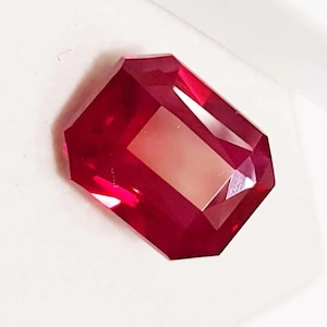 Peut inclure: Une pierre précieuse rubis rectangulaire et facettée, d'un rouge profond. La gemme présente une surface polie avec de multiples facettes qui réfléchissent la lumière, créant un aspect brillant. La pierre est posée sur un fond blanc.