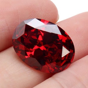 Könnte beinhalten: Ein facettierter, ovalförmiger, tiefroter Edelstein. Der Edelstein hat mehrere reflektierende Oberflächen, die das Licht einfangen und ein brillantes Schauspiel erzeugen. Der Stein wird in einer Hand gehalten, um seine Größe und Farbe zu zeigen.
