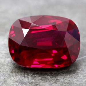 Könnte beinhalten: Ein tiefroter Rubin-Edelstein mit einem Kissen-Schliff. Der Edelstein ist facettiert und hat einen hohen Glanz.