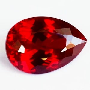 May include: A pear-shaped faceted gemstone with a deep, vibrant red color. The stone features a polished surface with multiple geometric facets that reflect light, creating bright highlights and dark shadows across its translucent, saturated body.