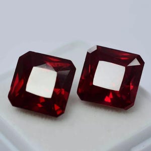 Könnte beinhalten: Zwei quadratisch geschliffene, tiefrote Edelsteine mit einem hellen, reflektierenden Zentrum. Die Edelsteine sind vor einem weißen Hintergrund platziert und zeigen ihre facettierten Oberflächen und ihre satte Farbe. Diese Edelsteine sind wahrscheinlich für die Schmuckherstellung.