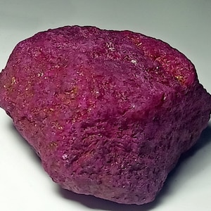 May include: A large, rough, pink ruby gemstone. The ruby has a deep, rich colour and a textured surface.