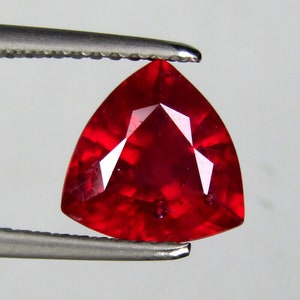 May include: A vibrant red triangular gemstone with a faceted cut. The gemstone is held by a pair of tweezers.
