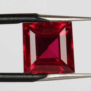 Könnte beinhalten: Ein quadratischer Edelstein in tiefroter Farbe mit einem facettierten Schliff. Der Edelstein wird von einer Pinzette gehalten.
