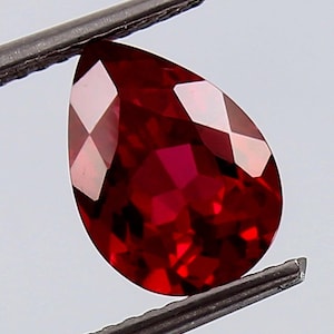 May include: A deep red pear-shaped gemstone with a faceted cut. The gemstone is held in a pair of tweezers.