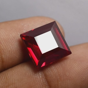 May include: A square-cut, deep red gemstone with a high degree of clarity. The gem's facets reflect light, creating bright highlights. The gemstone is held in a hand, suggesting its size and showcasing its colour.