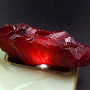 Peut inclure: Une grosse pierre précieuse de rubis taillée grossièrement, de couleur rouge foncé. Le rubis est éclairé par le bas, ce qui met en valeur ses facettes et sa translucidité.
