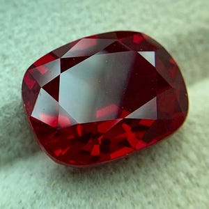 May include: A cushion-cut gemstone with a deep, saturated red color and a faceted surface that creates geometric light reflections. The stone has a rounded rectangular shape with polished edges, showcasing a clear, transparent, and vibrant red hue.