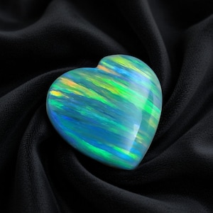 Puede incluir: Una piedra preciosa de ópalo en forma de corazón con una vibrante exhibición de colores. El ópalo presenta patrones arremolinados de tonos azules, verdes y amarillos. La gema se encuentra sobre un fondo negro texturizado, resaltando sus cualidades iridiscentes.