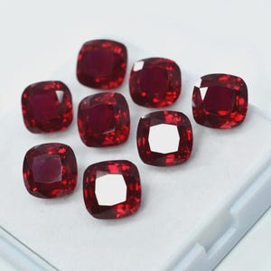 Peut inclure: Huit pierres précieuses rouges, taillées en coussin, sont disposées sur une surface blanche. Les gemmes ont une forme carrée aux coins arrondis et une surface brillante et réfléchissante. La couleur est un rouge riche et saturé, avec des tons plus foncés.