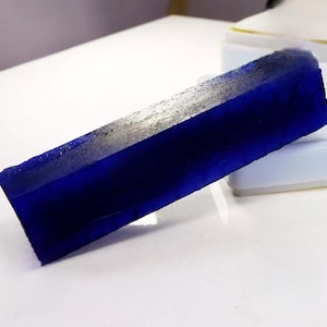 May include: A striking, rectangular, deep blue gemstone with a faceted, angular design. The gem has a polished, reflective surface and a raw, textured edge. The colour is a rich, saturated blue, with hints of lighter tones along the edges.