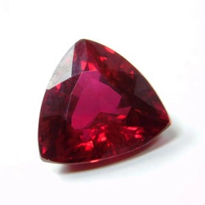 Peut inclure: Une pierre précieuse taillée en forme triangulaire, d'un rouge profond. La surface polie reflète la lumière, créant un éclat brillant. La gemme est posée sur un fond blanc.