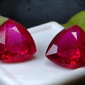 Peut inclure: Deux rubis triangulaires facettés. Les pierres précieuses sont d'un rouge vif avec un haut degré de clarté, reflétant la lumière. Elles sont présentées sur une surface blanche, avec une feuille verte floue en arrière-plan.