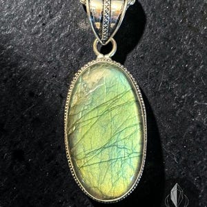May include: An oval labradorite pendant set in a silver frame with a rope-style bail, suspended from a silver rope chain. The stone displays a green and yellow iridescent sheen with natural internal fractures. The logo Ritualis is visible.