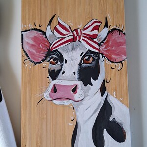 Peut inclure: Planche à découper en bois peinte à la main représentant une vache avec un bandeau à nœud rayé rouge et blanc. La vache a des marques noires et blanches, des oreilles et un nez roses, et des yeux bruns.
