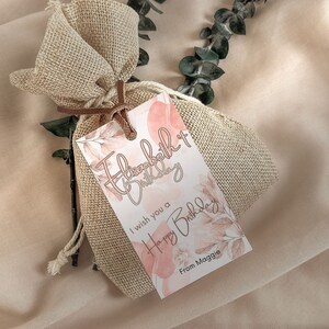 May include: A white gift tag with pink watercolor floral designs and the text "Happy Birthday" and "I wish you a Happy Birthday from Maggie". The tag is tied to a small burlap bag.